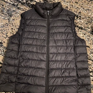 Svart dunväst med dragkedja - En stilren svart dunväst med horisontella sömmar och hög krage. Västen har en hel dragkedja framtill och är perfekt att slänga över en hoodie eller tröja för extra värme. Enkel och clean design som funkar till det mesta.