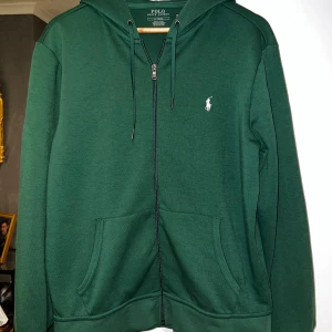 Grön hoodie från Polo Ralph Lauren - Mörkgrön hoodie från Polo Ralph Lauren med dragkedja, huva och snörning. Klassisk vit logga broderad på bröstet och två stora fickor framtill. Perfekt för chill dagar och enkel att matcha med jeans eller joggers.