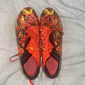 Adidas Messi fotbollsskor i rött och gult - Säljer ett par Adidas Messi fotbollsskor med grymt grafiskt mönster i rött, gult och svart. Skorna har snörning, låg profil och texten '360 Agility' på sidan. Perfekta för dig som vill sticka ut på planen och få max grepp med dobbarna. Högsta moddelen