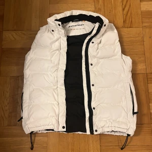 Vit dunväst från Everest, strl 38 - Snygg vit dunväst från Everest med svart foder och dragkedja framtill. Västen har hög krage, justerbar dragsko nertill och praktiska fickor med dragkedja. Perfekt för lager-på-lager och passar till många olika outfits.