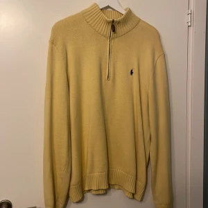 Gul stickad half zip från Polo Ralph Lauren - Snygg gul stickad tröja från Polo Ralph Lauren med half zip och hög krage. Tröjan har broderad logga på bröstet och är tillverkad i 100% bomull. Perfekt för dig som gillar klassisk och avslappnad stil.