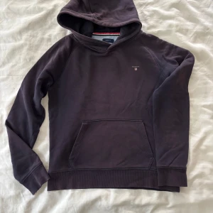 Mörklila hoodie från Gant - Snygg mörklila hoodie från Gant med klassisk huva och magficka. Tröjan har långa ärmar, ribbade muddar och en liten Gant-logga broderad på bröstet. Perfekt för en chill och avslappnad stil. 13-14 år. Skriv för att se deffekt på armen 