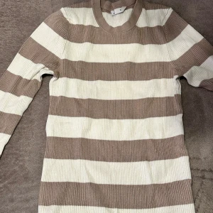 Randig ribbad tröja i beige och vitt - Säljer en ribbad långärmad tröja med breda beige och vita ränder. Tröjan har rund halsringning och är figurnära i modellen, perfekt för dig som gillar en stilren och enkel look.