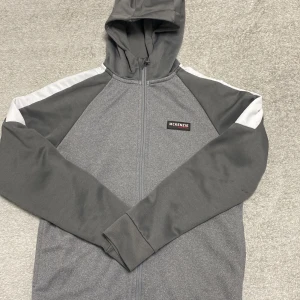 Grå hoodie från McKenzie med huva - Snygg grå hoodie från McKenzie med hel dragkedja och huva. Tröjan har vita detaljer längs axlarna och ärmarna, samt ett stilrent McKenzie-märke på bröstet. Mjuk insida och långärmad design, perfekt för chill dagar eller träning.