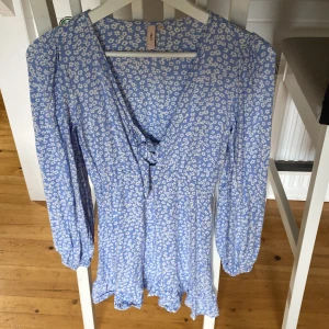 Blommig blå kortklänning från Nelly - Supersöt blå kortklänning från Nelly med vitt blommigt mönster. Klänningen har v-ringning med snörning framtill, långa puffärmar med resår vid ärmslut och volangkant nedtill. Perfekt för vår och sommar, och materialet känns lätt och skönt.