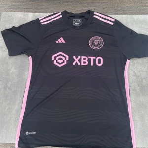 Inter Miami svart fotbollströja Adidas L - Snygg svart Inter Miami fotbollströja från Adidas i storlek L. Tröjan har rosa detaljer, klubbmärke och sponsortryck framtill samt rosa ränder på axlarna. Tillverkad i lätt och ventilerande material, perfekt för match eller träning.