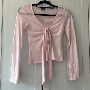 Ljusrosa rosetttopp från Gina Tricot - Supersöt ljusrosa omlottopp från Gina Tricot med lång ärm och knyt framtill. Toppen har v-ringning och croppad passform, perfekt för att skapa en feminin look. Mjuk och stretchig i materialet, passar till både jeans och kjol.