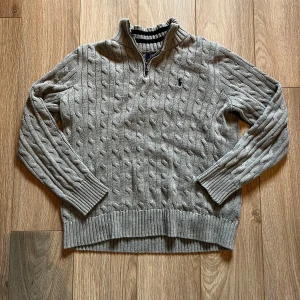 Grå stickad half zip tröja Polo Ralph Lauren - Säljer en grå stickad tröja från Polo Ralph Lauren med half zip och kabelstickat mönster. Tröjan har långärmade ribbade muddar och krage, samt den klassiska broderade loggan på bröstet. Perfekt för dig som gillar stilren och chill look. Storleken är L men skulle säga att den är mer Medium