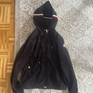 Svart vindjacka från Moncler med huva - Svart vindjacka från Moncler med huva och dragkedja framtill. Jackan har Moncler-logga på ärmen, snörning i huvan och en snygg rand i rött, vitt och blått på huvkanten. Tillverkad i lätt och glansig polyester, perfekt för blåsiga dagar.