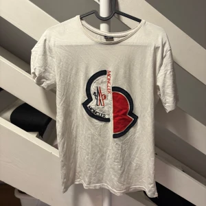 Vit Moncler t-shirt med stor logga - Vit t-shirt från Moncler i bomull med stor broderad logga i svart och rött på bröstet samt mindre logotyp-patch på ärmen. Klassisk rund halsringning och korta ärmar. Perfekt för dig som gillar streetwear och vill sticka ut med en ikonisk design.