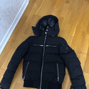Svart dunjacka Moose Knuckles - Snygg svart dunjacka från Moose Knuckles med huva och flera dragkedjefickor. Jackan har quiltad design, ribbade muddar och coola detaljer på ärmen. Perfekt för kalla dagar och ger en riktigt clean look. Lite skador men vid snabb affär så kan vi lösa ett bra pris. Kom privat om du vill ha fler bilder på jackan.