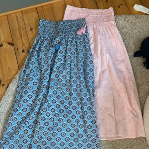 Två långa kjolar med smock och tofsar - Säljer två långa kjolar med smock i midjan och dekorativa tofsar. En är ljusrosa med diskret mönster, den andra blå med grafiskt mönster i vitt och svart. Båda är vida och luftiga, perfekta för varma dagar. Materialet känns som bomull och de har en avslappnad passform.