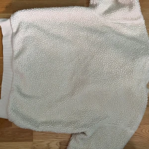 Vit teddy hoodie från GANT, M - Supermjuk och fluffig vit teddy hoodie från GANT med broderad SPORTSWEAR-text och logga på bröstet. Tröjan har huva med snörning, ribbade muddar och känguruficka. Perfekt för kalla dagar när du vill vara både varm och snygg.