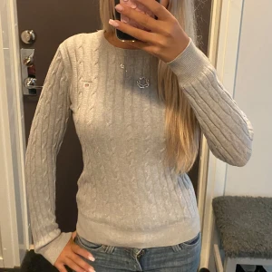 Ljusgrå kabelstickad tröja från Gant - Supersnygg ljusgrå kabelstickad tröja från Gant i stretchig bomull. Klassisk rund halsringning och diskret logga på bröstet. Perfekt till jeans eller kjol för en clean och stilren look. Ordinarie pris ca 1000kr💗