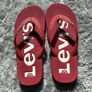 Röda flip-flops från Levi's - Snygga röda flip-flops från Levi's med stort vitt logotryck på sulan och matchande röd rem med logga. Tillverkade i syntetmaterial med mönstrad sula för extra grepp. Perfekta för sommardagar och strandhäng.