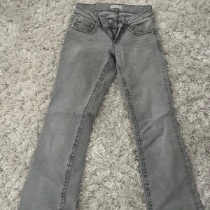 Grå bootcut jeans från Gina Tricot - Snygga grå jeans från Gina Tricot med bootcut passform. Jeansen har låg midja, coola fickdetaljer med knappar bak och fram, samt klassisk femficksdesign. Perfekta för dig som gillar en avslappnad men trendig look.