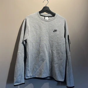 Grå Nike sweatshirt med dragkedja storlek S - Snygg grå sweatshirt från Nike med svart logga på bröstet och cool detalj med svart dragkedja på ärmen. Tröjan har rund halsringning och långa ärmar. Perfekt för dig som gillar sportig och clean stil. Helt oanvänd nypris 1099