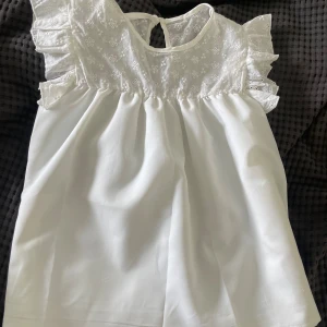 Vit blus med broderade volangärmar - Supersöt vit blus med broderade blommor upptill och volangärmar. Blusen har en luftig passform och knäppning i nacken. Perfekt för en fräsch och romantisk look under varma dagar. Säljer för jag aldrig andvänt blusen