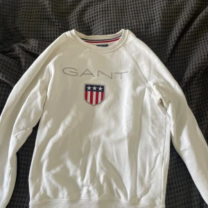 Vit långärmad tröja från GANT - Vit långärmad tröja från GANT i storlek S. Tröjan har ett broderat GANT-logo och ett snyggt emblem med stjärnor och ränder i blått och rött på bröstet. Rund halsringning med diskret röd och marinblå rand på insidan. Mjuk bomullskvalitet, perfekt för chill dagar.