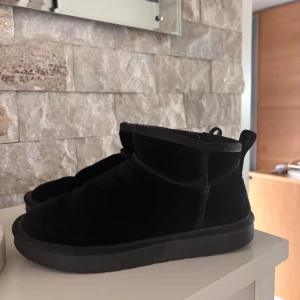 Svarta mockaboots med platt sula - Svarta boots i mjuk mocka med platt sula och rund tå. Skorna har en enkel och stilren design som passar perfekt till höst och vinter. Bekväma och lätta att matcha med olika outfits. (Uggs)