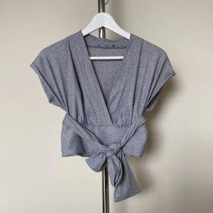 Glittrig silver omlottopp med knyt - Snygg glittrig silverfärgad omlottopp med djup v-ringning och breda axlar. Toppen har ett brett knytband i midjan som ger en cool och trendig look. Perfekt för dig som vill sticka ut med lite extra shine.