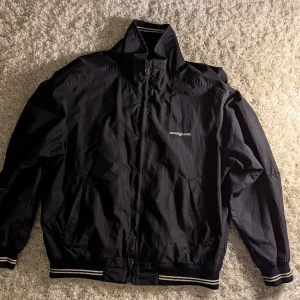 Vintage Henri Lloyd Jacka M - Svart vindjacka från Henri Lloyd med broderad logga på bröstet och ryggen. Jackan har dragkedja, ribbade muddar med vita ränder och rutigt innerfoder. Perfekt för dig som gillar klassisk stil med sportig touch.