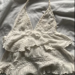 Vit omlott topp med volanger från Nelly - Supersöt vit omlott topp från Nelly i storlek XS. Toppen har halterneck, volangdetaljer och smockad midja. Materialet är mjukt och lätt med diskreta ränder. Perfekt för sommarens alla häng! Snyggt att matcha med jeans eller kjol.