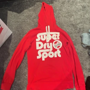 Röd Superdry Sport hoodie med tryck - Riktigt snygg röd hoodie från Superdry Sport med stort vitt tryck framtill och coola ränder i vitt och svart på ryggen. Tröjan har huva med snörning och är tillverkad i ett sportigt, lätt material som andas. Perfekt för dig som gillar streetwear och sportig stil.