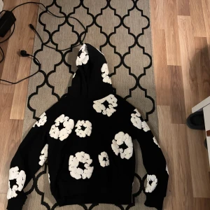 Svart hoodie med vita blommor Denim Tears - Svart hoodie från Denim Tears i storlek S med stora vita blommor tryckta över hela tröjan. Riktigt tung tröja som passar riktigt bra till allt och alla!Tröjan har huva och långa ärmar, perfekt till dig som vill sticka ut och se riktigt snygg ut, 