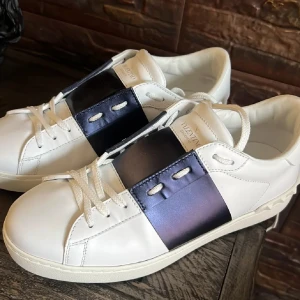 Valentino Garavani Open  - Säljer ett par Valentino Garavani Open sneakers i vitt skinn med en bred marinblå läderpanel över mitten. Skorna har vita snören, rund tå och platt sula med coola nitar på hälen. Perfekt för dig som gillar lyxiga och stilrena sneakers med en unik touch. Liten defekt vid ena remmen 