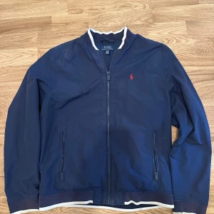 Marinblå vindjacka från Polo Ralph Lauren - Snygg marinblå vindjacka från Polo Ralph Lauren med vit ribbad krage och nederkant. Jackan har dragkedja framtill, två fickor med zip och den klassiska röda Polo-loggan på bröstet. Materialet är en mix av bomull och nylon med polyesterfoder. Perfekt för chilliga dagar.