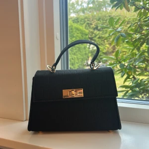 Svart handväska med krokoprägling - Stilren svart handväska med krokopräglat mönster och guldfärgade metalldetaljer. Väskan har både handtag och justerbar axelrem, samt snyggt vridlås framtill. Perfekt storlek för mobil, plånbok och andra essentials. Ger en classy vibe till din outfit.