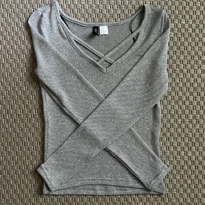 Glittrig silver topp från Divided XS - Snygg långärmad topp i silver från Divided. Toppen har glittrigt material, v-ringning med dekorativa band framtill och en tight passform. Perfekt för dig som vill sticka ut med lite extra shine. Superfin att styla med jeans eller kjol!