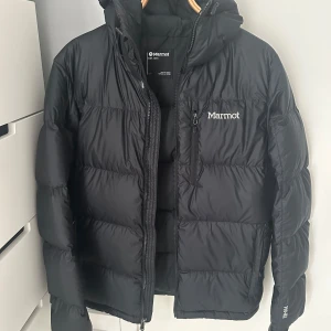 Svart pufferjacka från Marmot - Säljer en svart pufferjacka från Marmot med huva och dragkedja framtill. Jackan har en quiltad design, bröstficka med dragkedja och logga på bröstet. Perfekt för kalla dagar när du vill hålla värmen och ändå se clean ut. Jackan har en defekt och det är hängaren som är av.