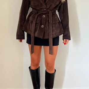 Brun äkta mockajacka med bälte - Snygg brun mockajacka med bälte i midjan och stora fickor framtill. Jackan har knappar och markerad midja för en trendig siluett. 