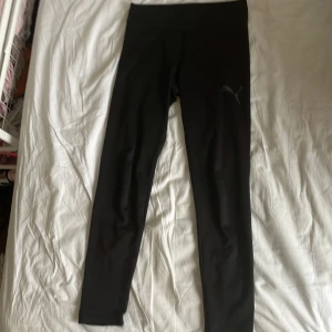Svarta leggings från Puma - Svarta leggings från Puma med klassisk logga på benet. De har en tight passform och är gjorda i ett stretchigt syntetmaterial som passar perfekt för träning. Låg midja och stilren design gör dem enkla att matcha med andra träningskläder.