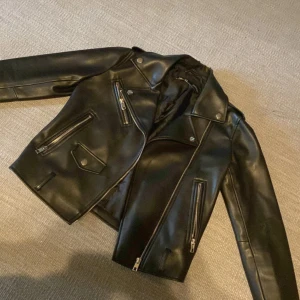 Svart skinnjacka från Zara - Säljer en svart snygg skinnjacka från Zara. Jackan har silvriga dragkedjor, knappar och detaljer, samt flera fickor framtill. Snygg passform och perfekt för dig som vill ha en edgy look.   Den är lite trasig i kragen (titta på bild 5), annars väldigt bra skick! 