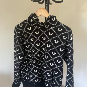 Svart True Religion hoodie med mönster - Svart hoodie från True Religion med helvitt logomönster och text i rutigt mönster över hela tröjan. Klassisk huva och långa ärmar, ribbade muddar och midja. Perfekt för dig som gillar streetwear och vill sticka ut med en fet logga.