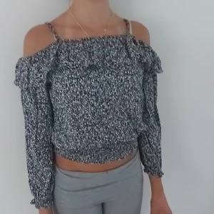 Svartvit blommig offshoulder blus - Supersöt svartvit blommig blus med offshoulder och smala axelband. Toppen har långa ärmar, volangdetalj upptill och smockad nederkant som ger en croppad känsla. Perfekt för dig som gillar trendiga och luftiga plagg med lite extra detaljer.