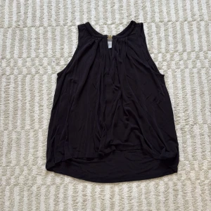 Svart ärmlös linnetopp med guldknäppe - Svart flowy linnetopp från H&M i storlek M. Toppen är ärmlös med snygga veck framtill och har ett coolt guldfärgat knäppe vid halsen, samt en öppning vid bröstkorgen. Perfekt för fest!