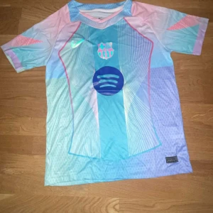 FC Barcelona x Nike Dri-Fit fotbollströja - Säljer en FC Barcelona x Nike Dri-Fit fotbollströja i pastellfärger med klubbmärke och Spotify-logga på bröstet. Tröjan är kortärmad och har ett unikt mönster i blått, rosa och lila. Tillverkad i lätt och ventilerande polyester för maximal komfort på planen.