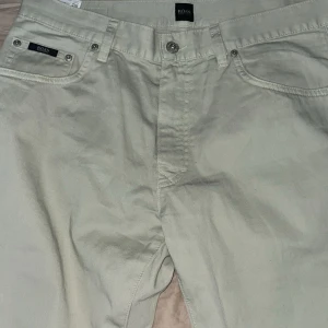 Beige chinos från BOSS, storlek 35/30 - Snygga beige chinos från BOSS med klassisk femficksdesign och diskret logga på fickan. Byxorna är raka i modellen och tillverkade i mjuk bomull som känns skön mot huden. Perfekta för en clean och stilren look.