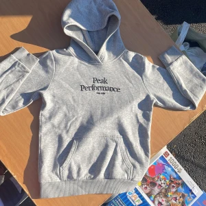 Grå hoodie Peak Performance 150 - Snygg ljusgrå hoodie från Peak Performance med broderad logga på bröstet. Klassisk känguruficka framtill, ribbade muddar och mjuk insida. Perfekt för chill dagar eller när du vill ha en clean och sportig look.