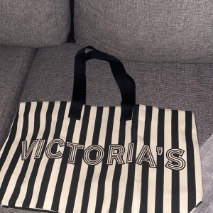 Randig shoppingväska Victoria's Secret - Stor och rymlig shoppingväska från Victoria's Secret med svartvita ränder och tydlig logga. Väskan har svarta handtag och insida i glansigt svart tyg. Perfekt för att bära med sig allt man behöver under dagen.