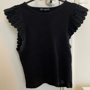 Svart ärmlös topp med volang från Zara - Supersnygg svart topp med broderade volangdetaljer på axlarna. Ärmlös modell i mjukt material, perfekt att styla med jeans eller kjol. Volangerna har en virkad look som ger extra edge till outfiten.