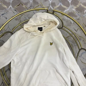 Vit hoodie från Lyle & Scott - Snygg vit hoodie från Lyle & Scott med klassisk gul logga på bröstet. Tröjan har huva med snörning och en stor magficka framtill. Perfekt för dig som gillar en clean och stilren look.