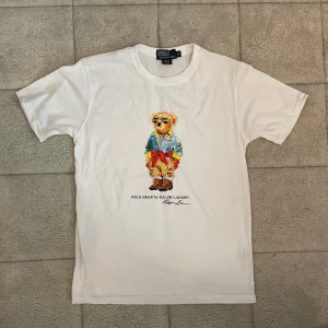 Vit Polo Bear t-shirt från Ralph Lauren - Vit t-shirt från Polo Ralph Lauren med ikoniskt Polo Bear-tryck på bröstet. Klassisk rund hals och korta ärmar. Trycket visar en björn i färgglada kläder och detaljerade accessoarer. Tillverkad i mjuk bomull för en skön känsla. Perfekt för dig som gillar streetstyle och preppy vibes. Passar även S