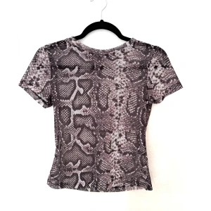 Ormmönstrad topp - Säljer en grå och svart topp med korta ärmar och coolt ormmönster. Toppen är figurnära och gjord i mjuk polyester som känns lätt mot huden. Perfekt för dig som vill sticka ut med ett edgy print.