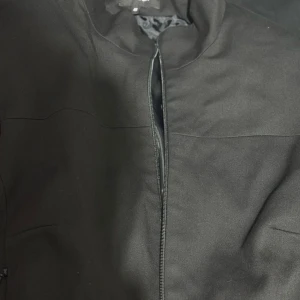 Svart bomberjacka från Lindex, stl 46 - Svart bomberjacka från Lindex i storlek 46. Jackan har minimalistisk design med dragkedja framtill och två fickor med dragkedja. Materialet är en mix av polyester, viskos och elastan, vilket ger en mjuk och följsam känsla. Perfekt för dig som gillar stilren och enkel look.