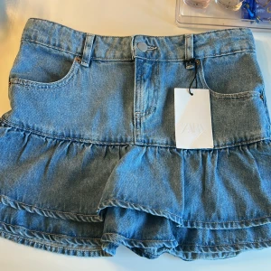 Volangkjol i jeans från Zara - Supersöt jeanskjol från Zara med volangdetaljer och klassisk femficksdesign. Kjolen är i ljusblå denim och har en kort, lekfull passform. Perfekt för dig som vill ha en trendig och avslappnad look med extra volym nertill.